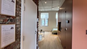 A hallway with a brick wall, a white door, a small round table with a plant, and a bed in the distance.