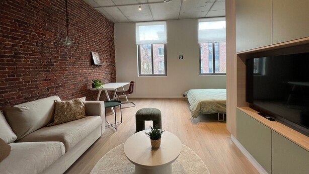 A modern living room with a brick wall, a sofa, a coffee table, a TV, and a bed.