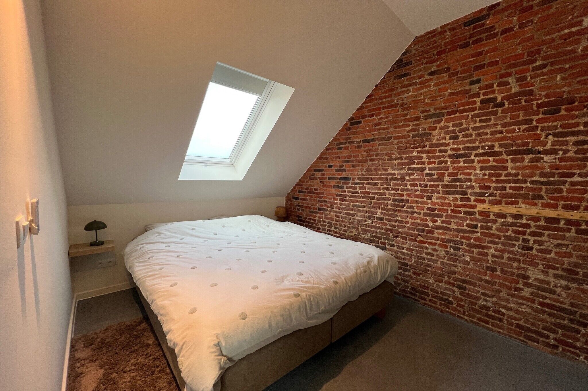 Quarto com teto inclinado, claraboia, parede de tijolos, cama com colcha branca e mesinha de cabeceira com abajur.