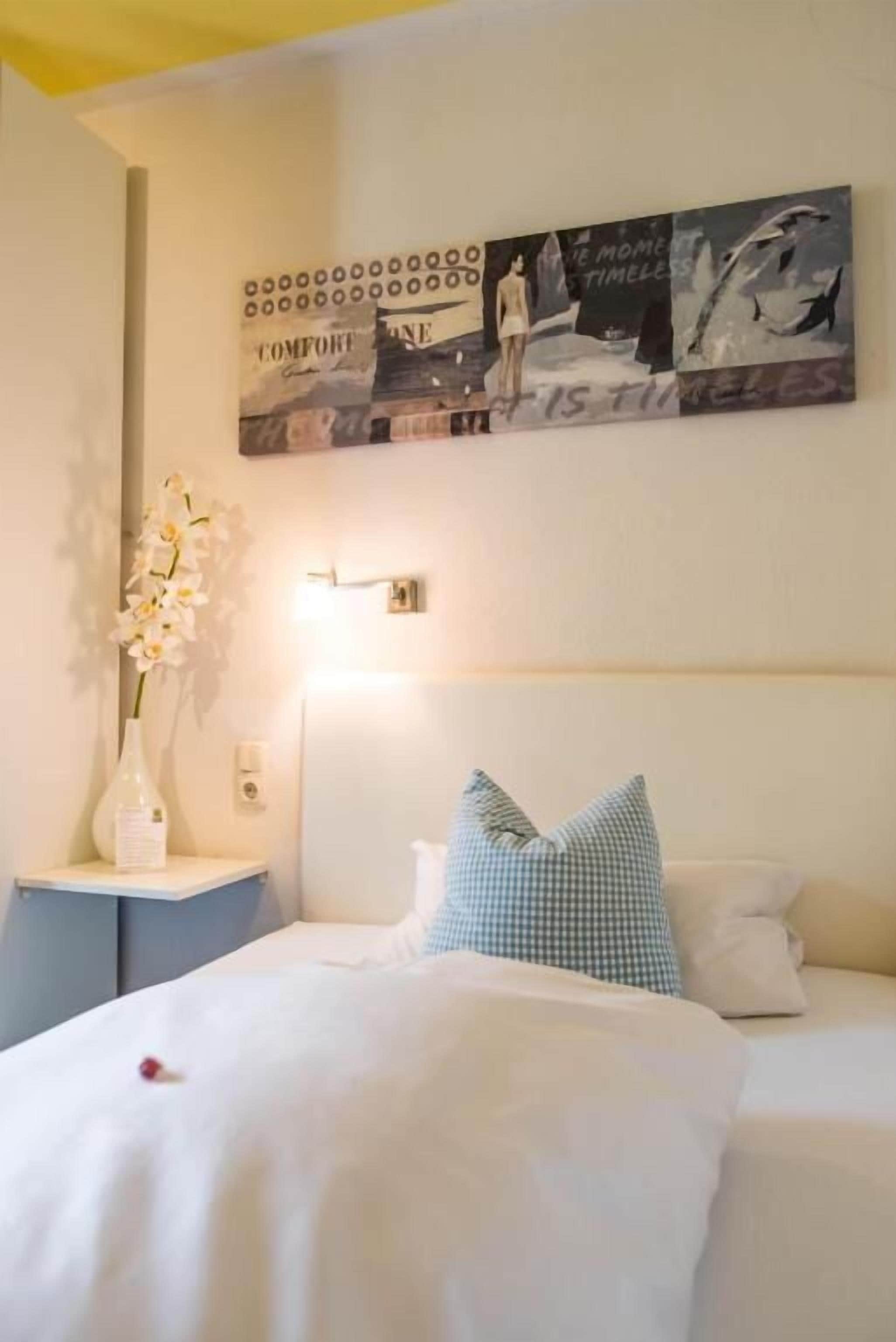 A bedroom with a bed, a nightstand, a vase with flowers, and a wall art piece.