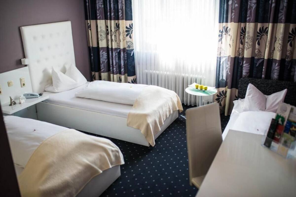 A hotel room with two beds, a small table with a lamp, and a window with curtains.