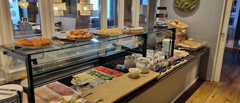 Un bufé con diversos alimentos, como pasteles, carnes y verduras, exhibidos sobre un mostrador de vidrio con utensilios para servir.