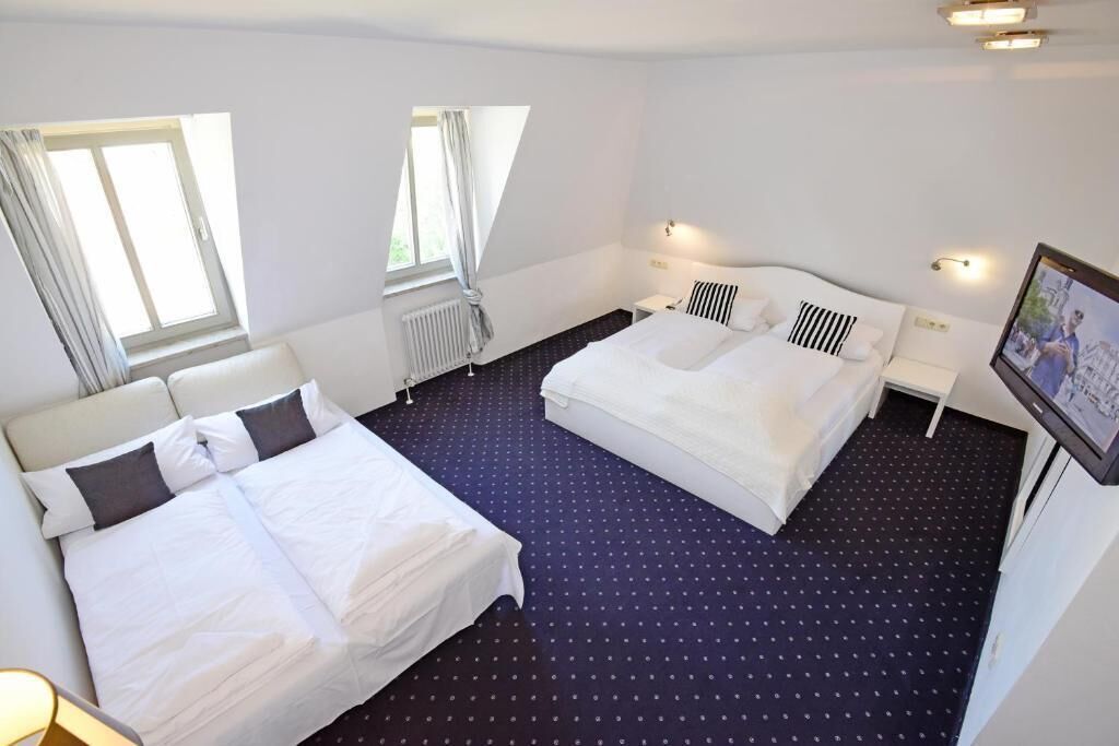 A hotel room with two beds, a window, a radiator, and a wall-mounted TV.