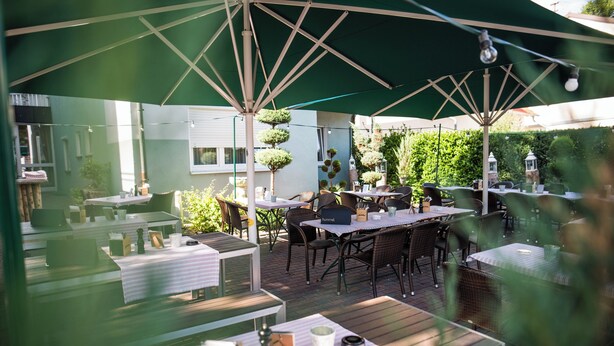 An outdoor patio area with tables and chairs under green umbrellas, surrounded by greenery and potted plants. - Hotel Domizil (Ingolstadt)