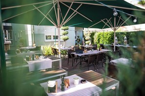 An outdoor patio area with tables and chairs under green umbrellas, surrounded by greenery and potted plants. - Hotel Domizil (Ingolstadt)