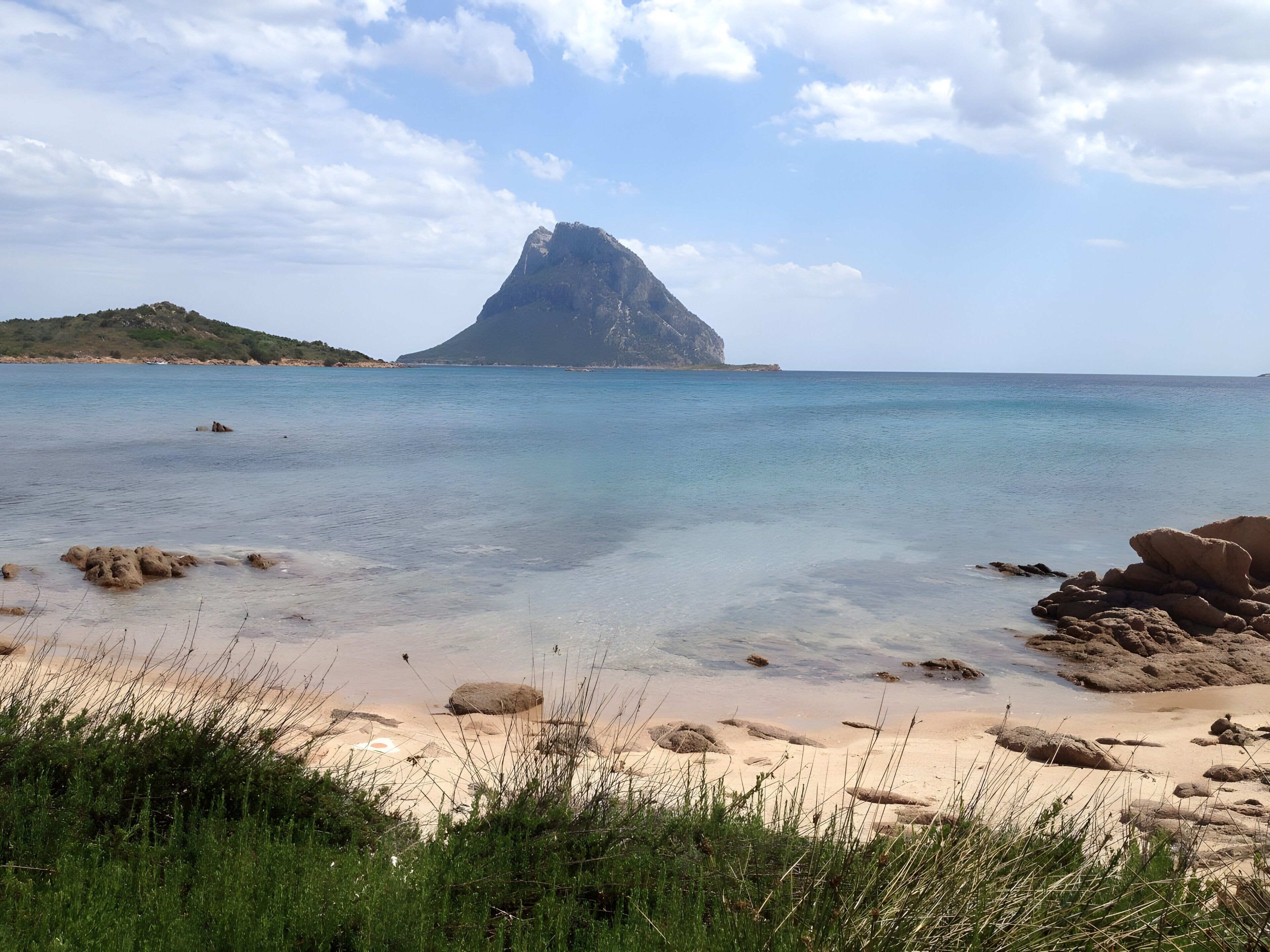 A beach with clear water, a large rock formation, and sparse vegetation.