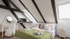 Ein Schlafzimmer mit schrägen Wänden, einem Bett mit einer grünen Tagesdecke, weißer Bettwäsche, einem Nachttisch mit Lampe und einem Spiegel auf einer Kommode.