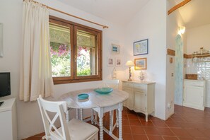 A dining area with a table, chairs, a window with curtains, a lamp, and kitchen cabinets. - Villetta Oleandro with Sea View, Private Terrace and Garden (San Teodoro)