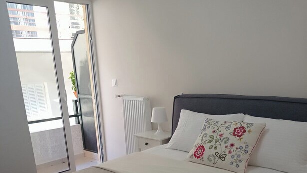 A neatly made bed with two folded towels, a bedside table with a lamp, and a window with a view of buildings. - Seizani Athens Stay: Cozy apartment in welcoming Kesariani (Kesariani, Attiki)