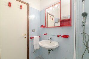 Casa de banho com lavatório branco, espelho com iluminação, chuveiro e suportes de toalha vermelhos.