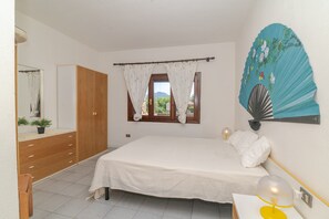 Um quarto com cama, guarda-roupa de madeira, janela com cortinas, mesa de cabeceira com abajur e ventoinha fixada na parede.