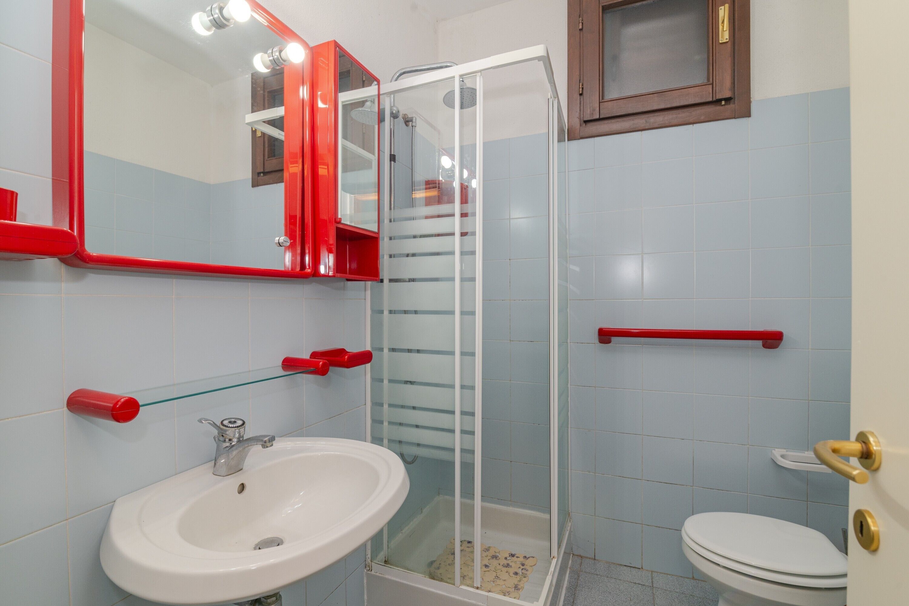 Un baño con lavamanos blanco, ducha con cerramiento de vidrio, inodoro y espejo con marco rojo.