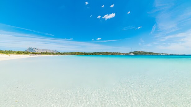 En strand med kristallklart turkost vatten, ett avlägset berg och en klar himmel.