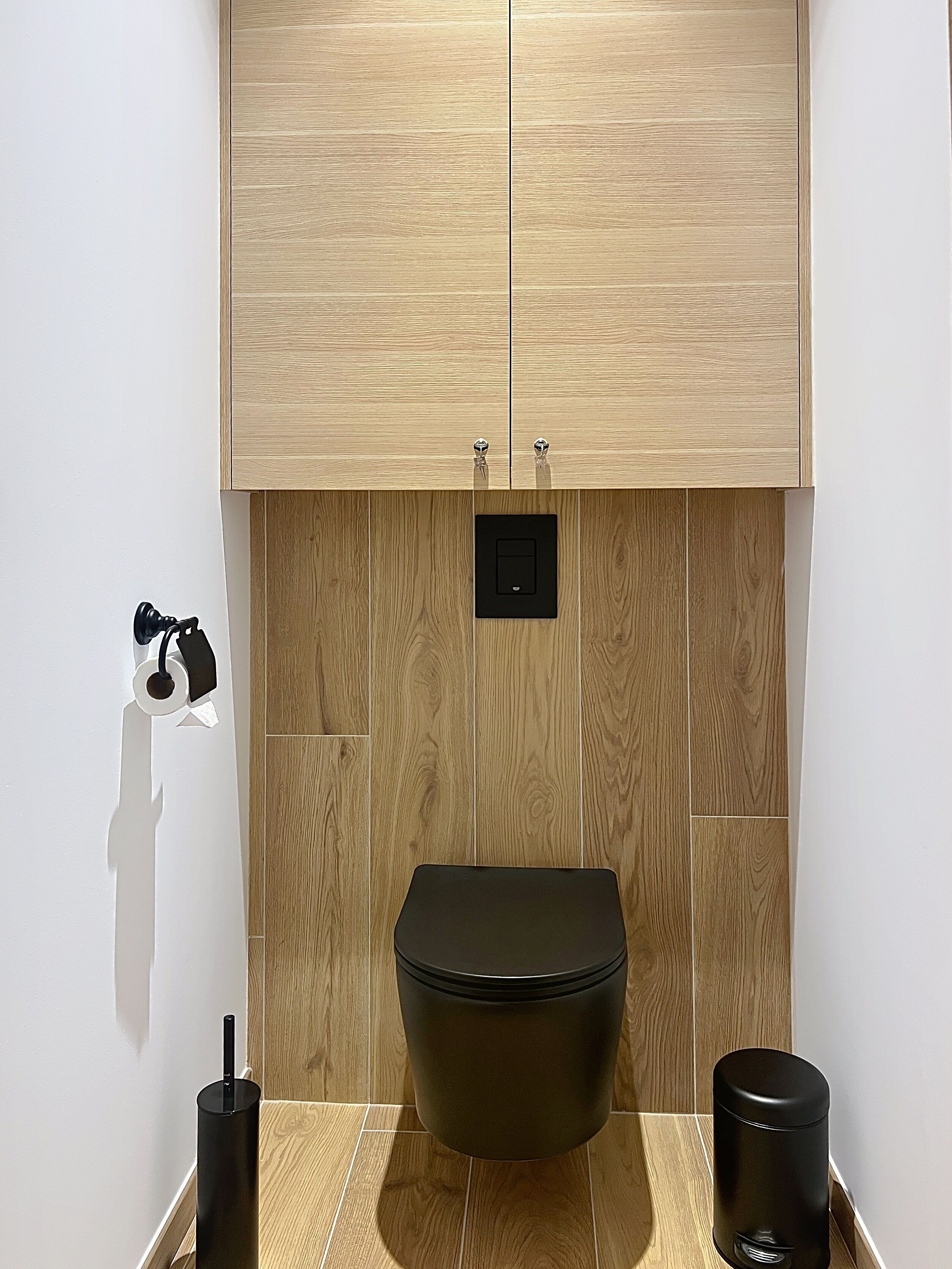 A bathroom with a toilet, a trash bin, a hand dryer, and a cabinet above the toilet.