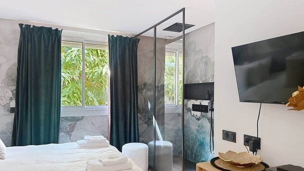 A modern hotel room with a bed, a shower area, a TV, and a view of greenery outside. - T2 Luxury Canet • Beach 50m - Romantic stay ❤️ Love room (Canet-en-Roussillon)