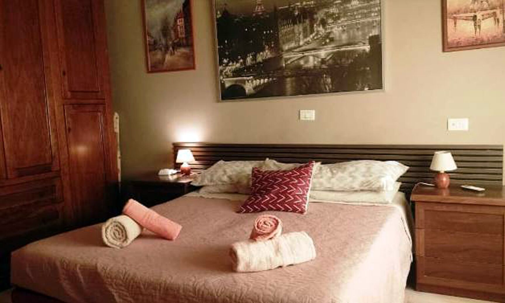 Een net opgemaakt bed met een roze sprei, twee kussens en opgerolde handdoeken. Twee nachtkastjes met lampen en een houten garderobe.
