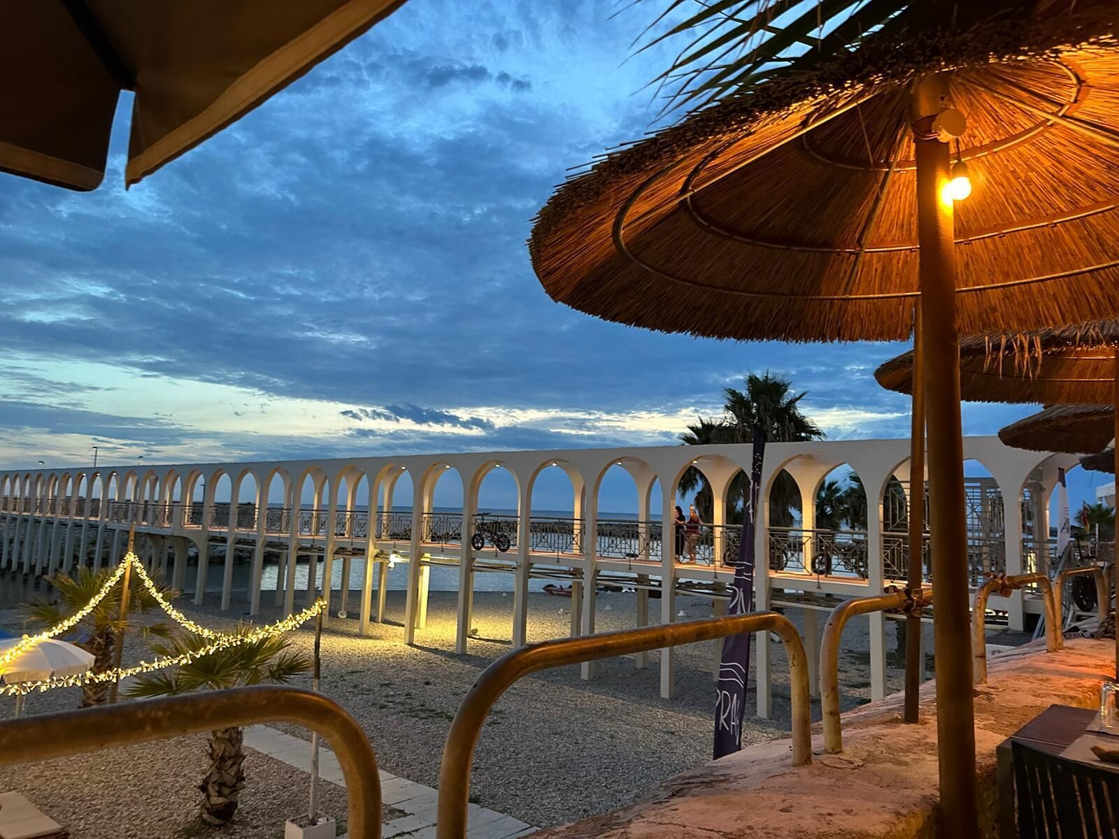 Une promenade en bord de mer agrémentée d’une série d’arcades, une plage bordée de palmiers et un parasol en chaume.