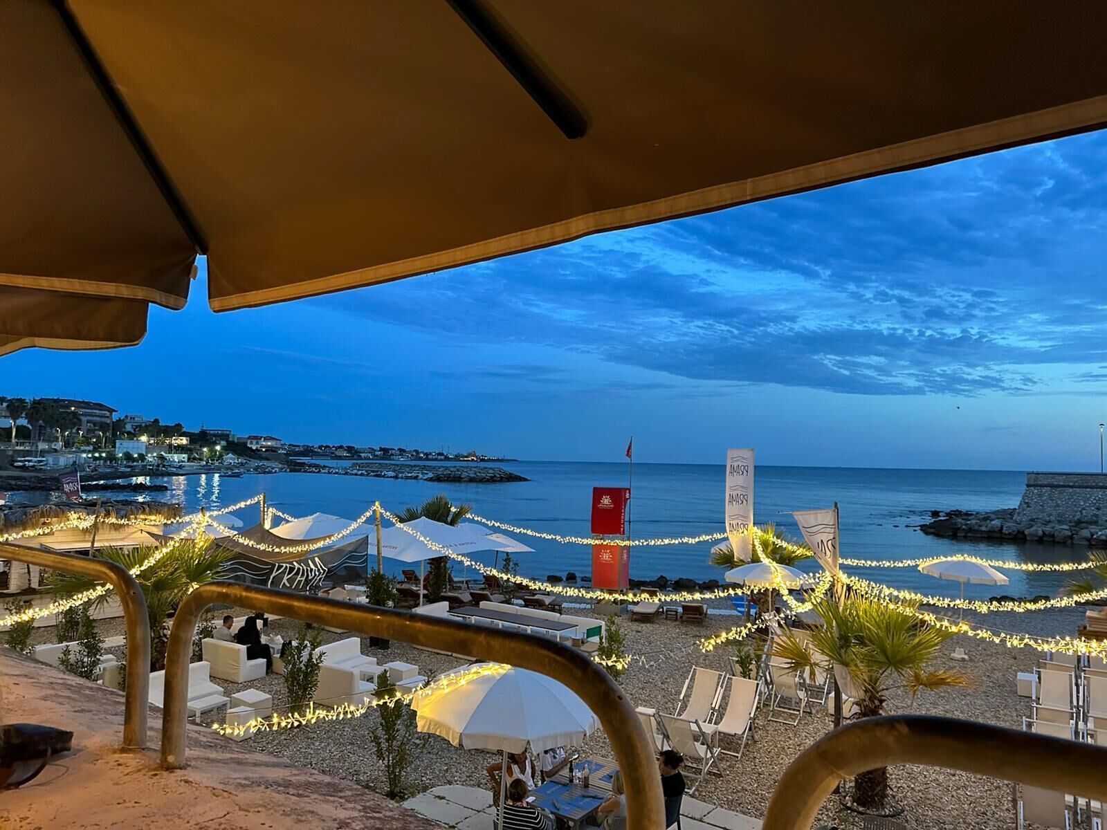 Ein Sitzbereich am Strand mit Sonnenschirmen und Lichterketten, mit Blick auf das Meer und eine ferne Küstenlinie.