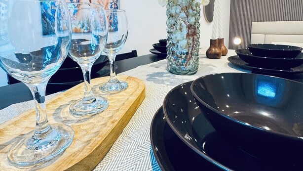Zona pranzo con tavolo moderno apparecchiato per due, con un bicchiere da vino in vetro, un bicchiere di vetro e un piatto nero. Sul tavolo c'è un vaso decorativo con fiori bianchi e sullo sfondo si intravede un quadro.