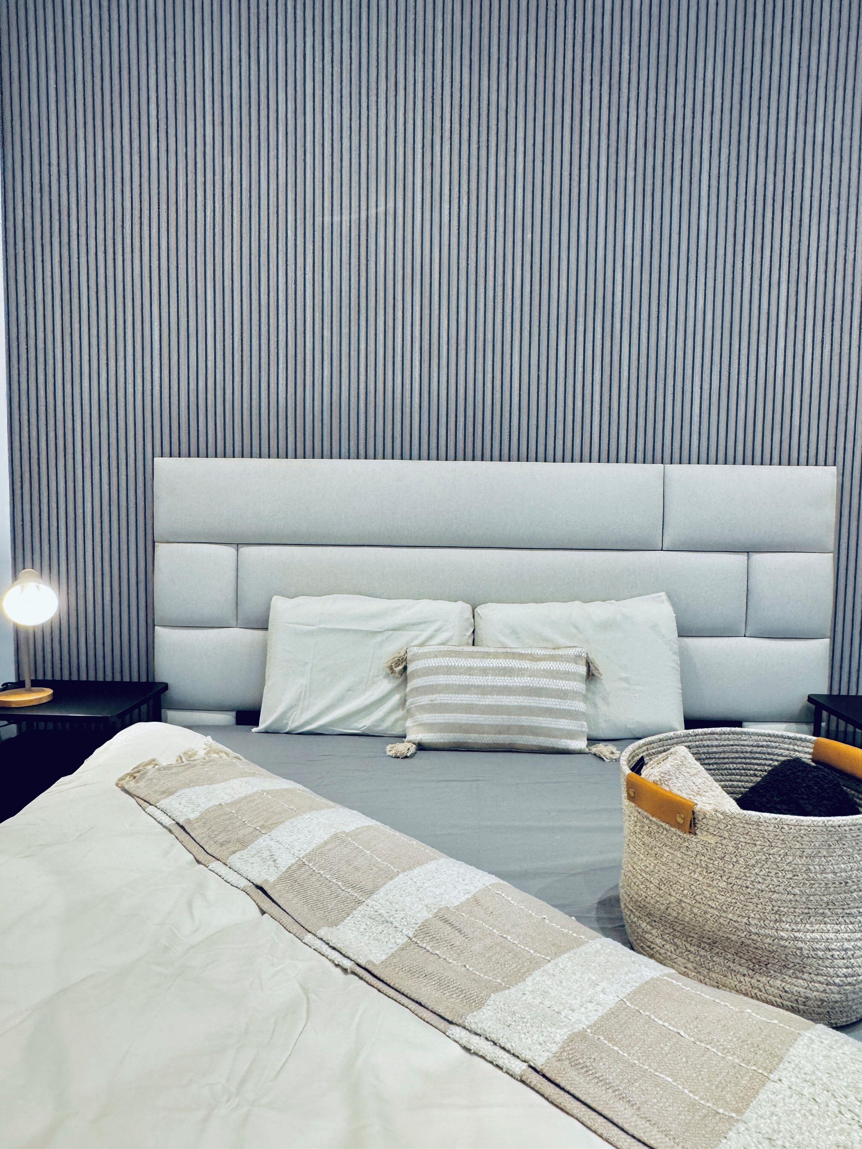 A modern bedroom with a striped wall, a large bed with multiple pillows, a bedside table with a lamp, and a woven basket on the floor.