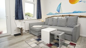 A modern living room with a grey sofa, a small white table, and a colorful rug. A large window with blue curtains is visible, and a sailboat painting adorns the wall. - Haus Concordia - Holiday apartment on the Mönchgut - Haus Concordia vacation apartment (Mönchgut)