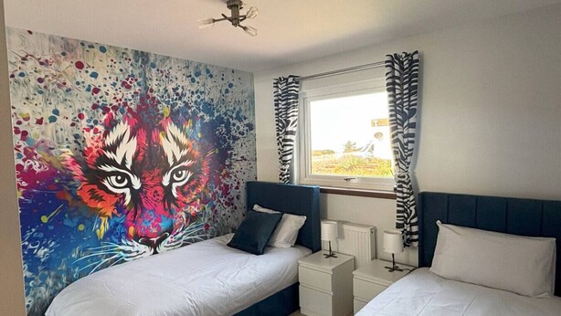 A bedroom with two beds, a colorful abstract painting on the wall, a window with curtains, and a ceiling fan. - The Burghead Hideaway – Calm & Comfort (Burghead)