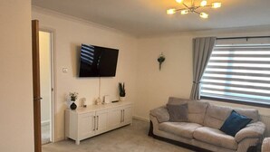 A living room with a beige sofa, a white cabinet, a flat-screen TV, and a window with blinds. - The Burghead Hideaway – Calm & Comfort (Burghead)