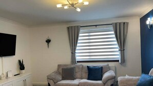 A living room with a sofa, a television mounted on the wall, and a window with curtains. - The Burghead Hideaway – Calm & Comfort (Burghead)