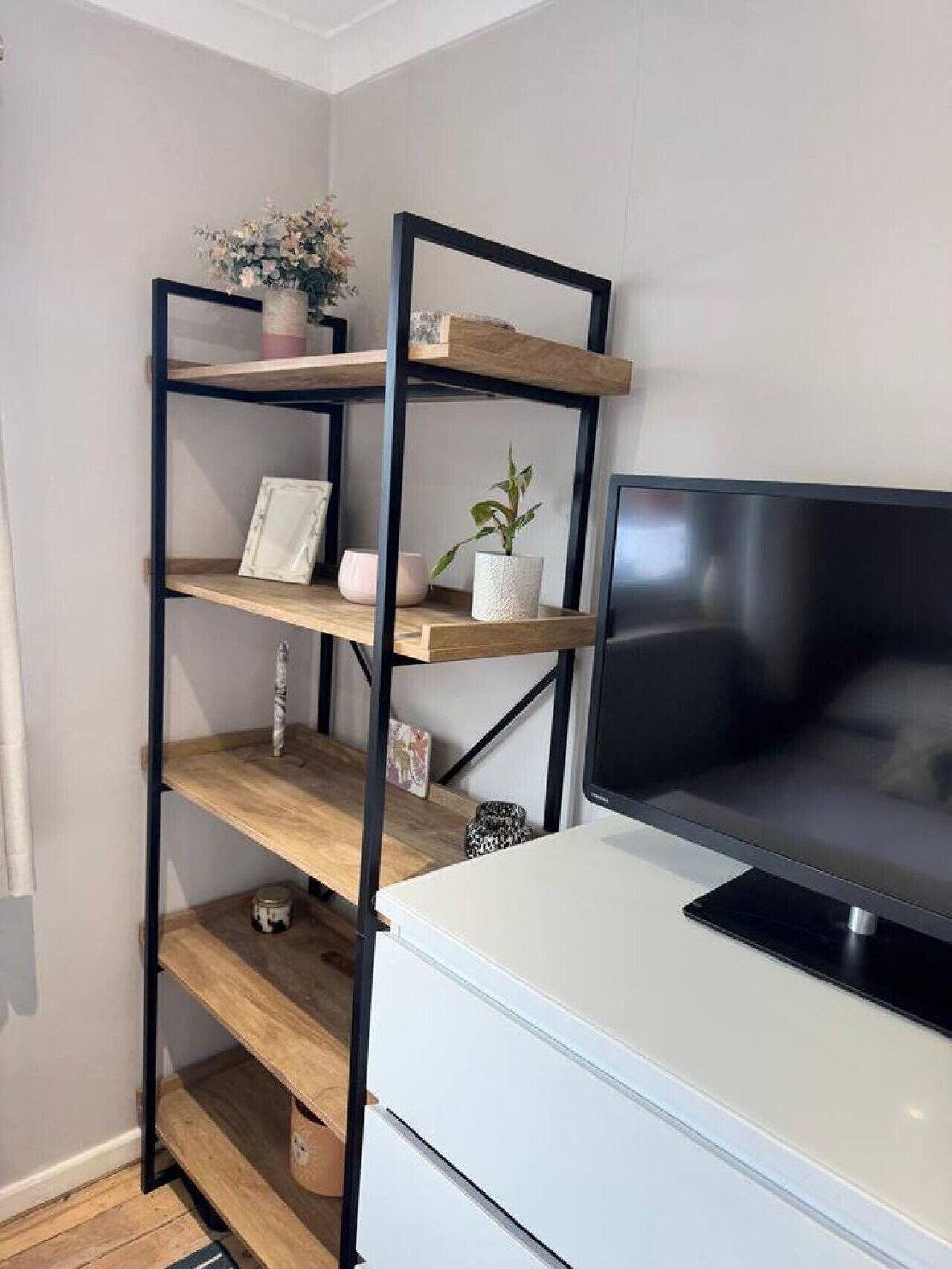 Ein Zimmer mit weißem Schreibtisch, einem schwarzen Fernseher und einer schwarzen Metallregaleinheit mit Holzböden.