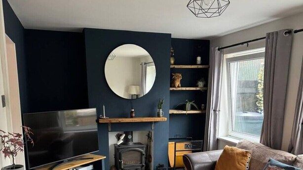 Una sala moderna con una pared de acento azul oscuro, un espejo redondo, un televisor de pantalla plana, un cofre de madera, un sofá con cojines y una chimenea.