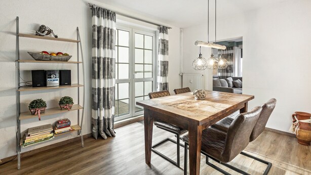 A dining area with a wooden table, chairs, a bookshelf, and a window with curtains. - Apartment 'Hoppernkopf' with Shared Terrace, Private Garden and Wi-Fi (Willingen (Upland))