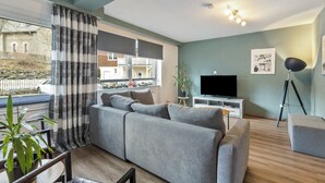 A modern living room with a grey sofa, a flat-screen TV, a floor lamp, and a large window with curtains. - Apartment 'Hoppernkopf' with Shared Terrace, Private Garden and Wi-Fi (Willingen (Upland))