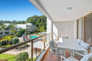 Ein Balkon mit weißem Tisch und Stühlen, der auf einen Swimmingpool und üppiges Grün blickt.