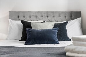 A bed with a tufted headboard, multiple pillows in various shades, and a dark throw blanket. - Foxden House – Your Home from Home in the Cotswolds. (Bourton-on-the-Water)