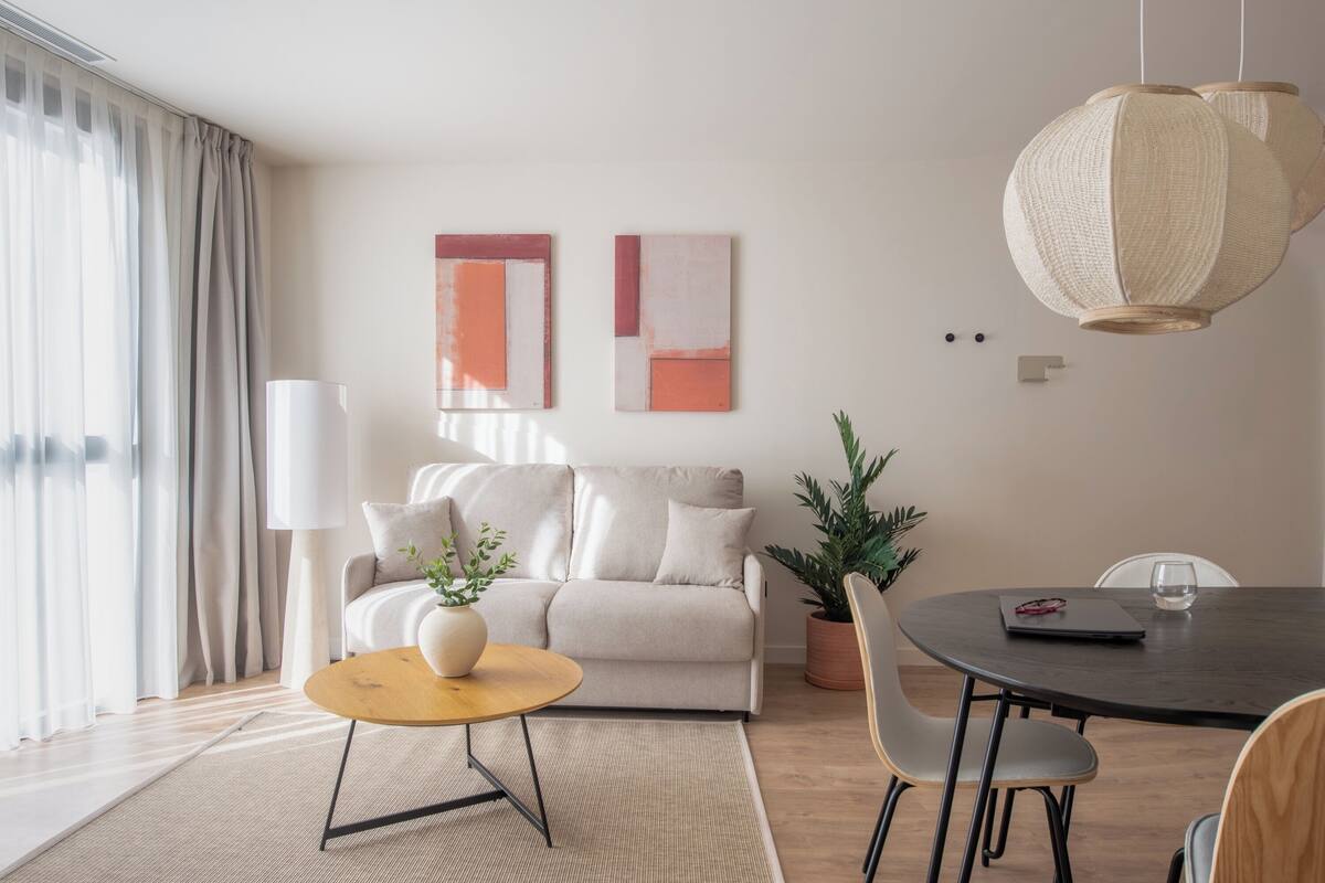 A modern living room with a sofa, round coffee table, and dining table. There are abstract paintings on the wall and a hanging light fixture.