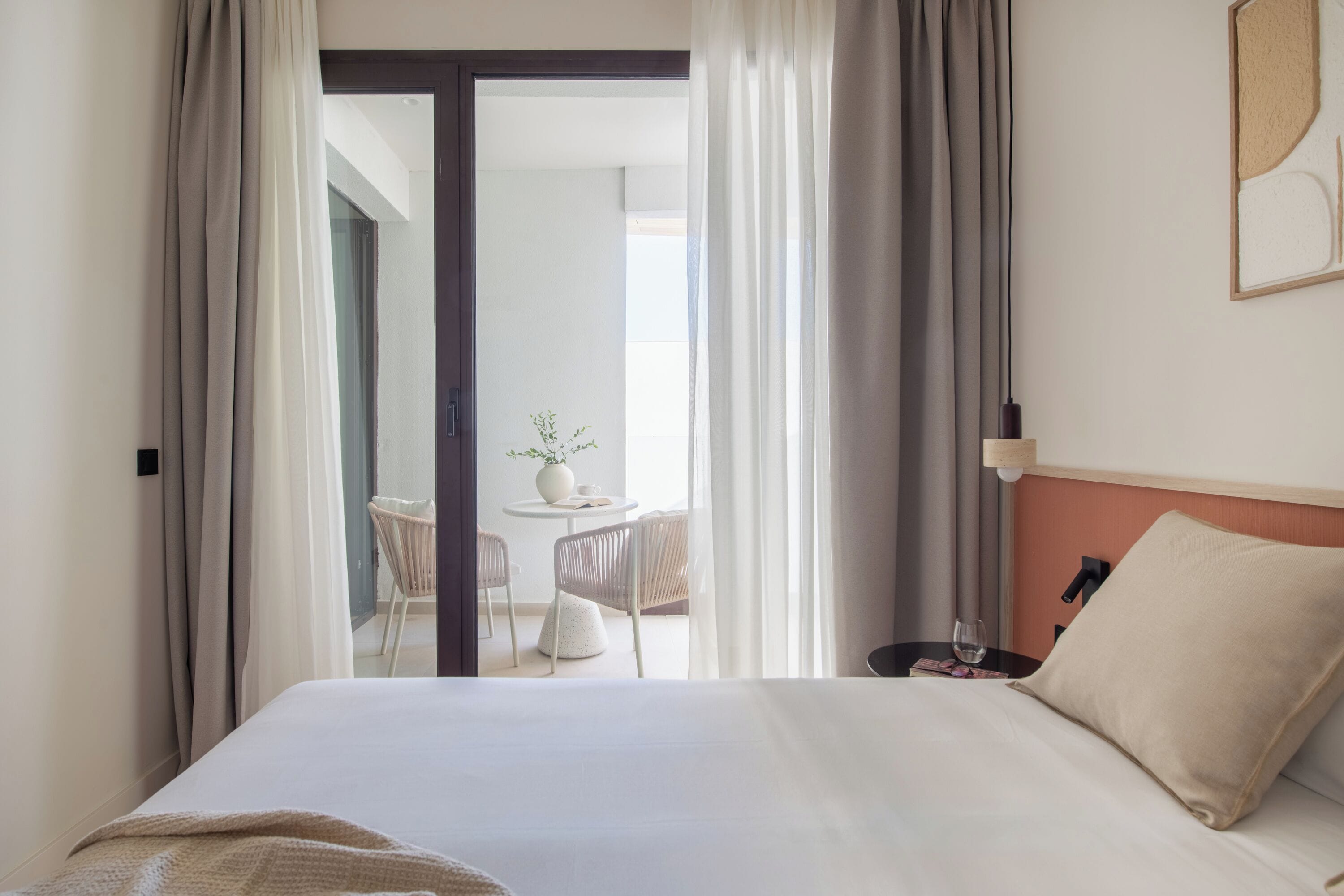 Habitación de hotel con cama, mesita de noche, lámpara y vistas a un balcón con mesa y sillas.