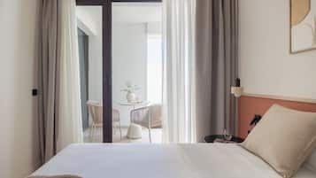 Habitación de hotel con cama, mesita de noche, lámpara y vistas a un balcón con mesa y sillas.