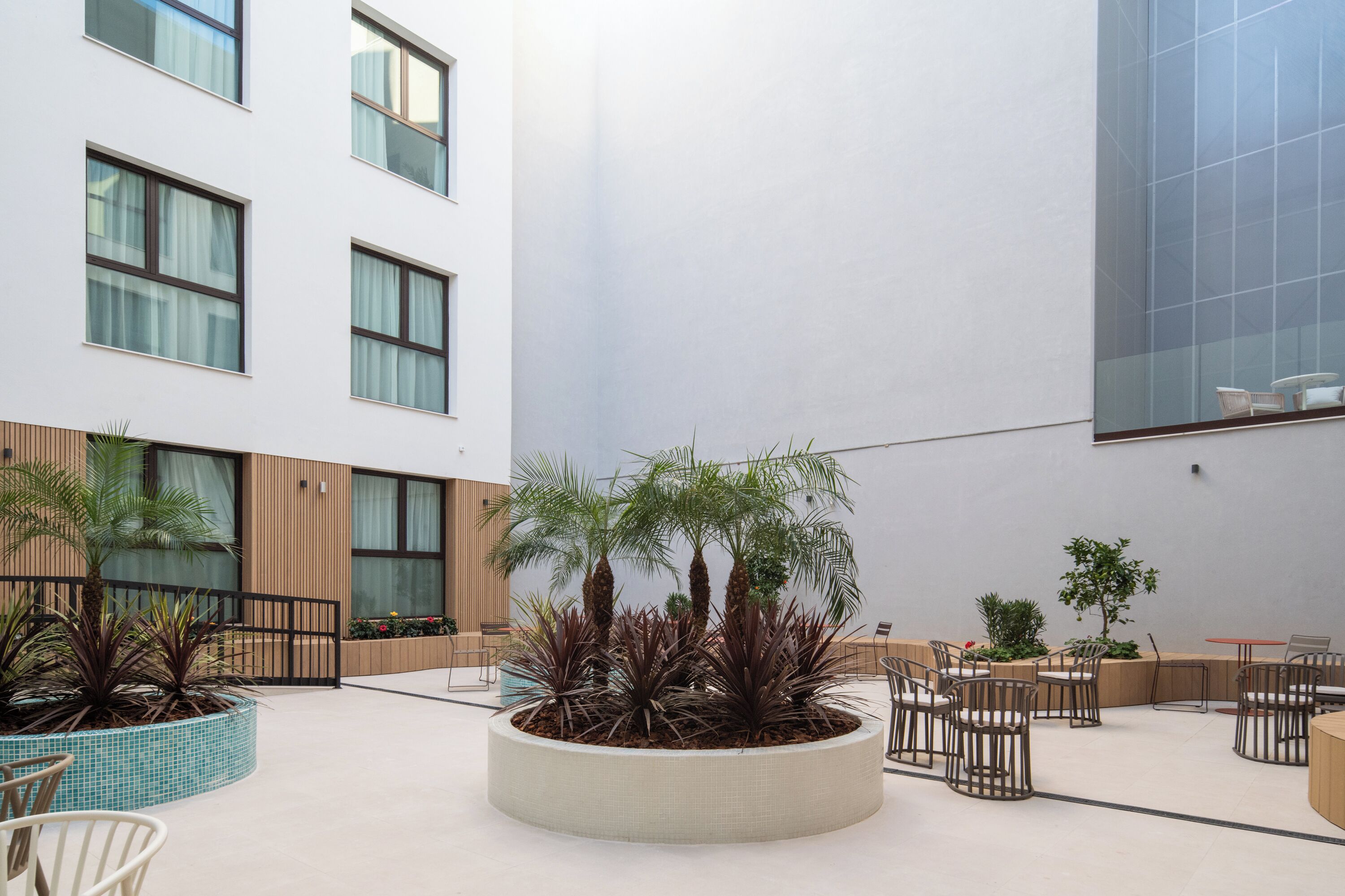 Un vaste patio intérieur aménagé avec des meubles modernes, un jardin central planté de végétaux et un bâtiment doté de grandes fenêtres et d’une façade en verre.