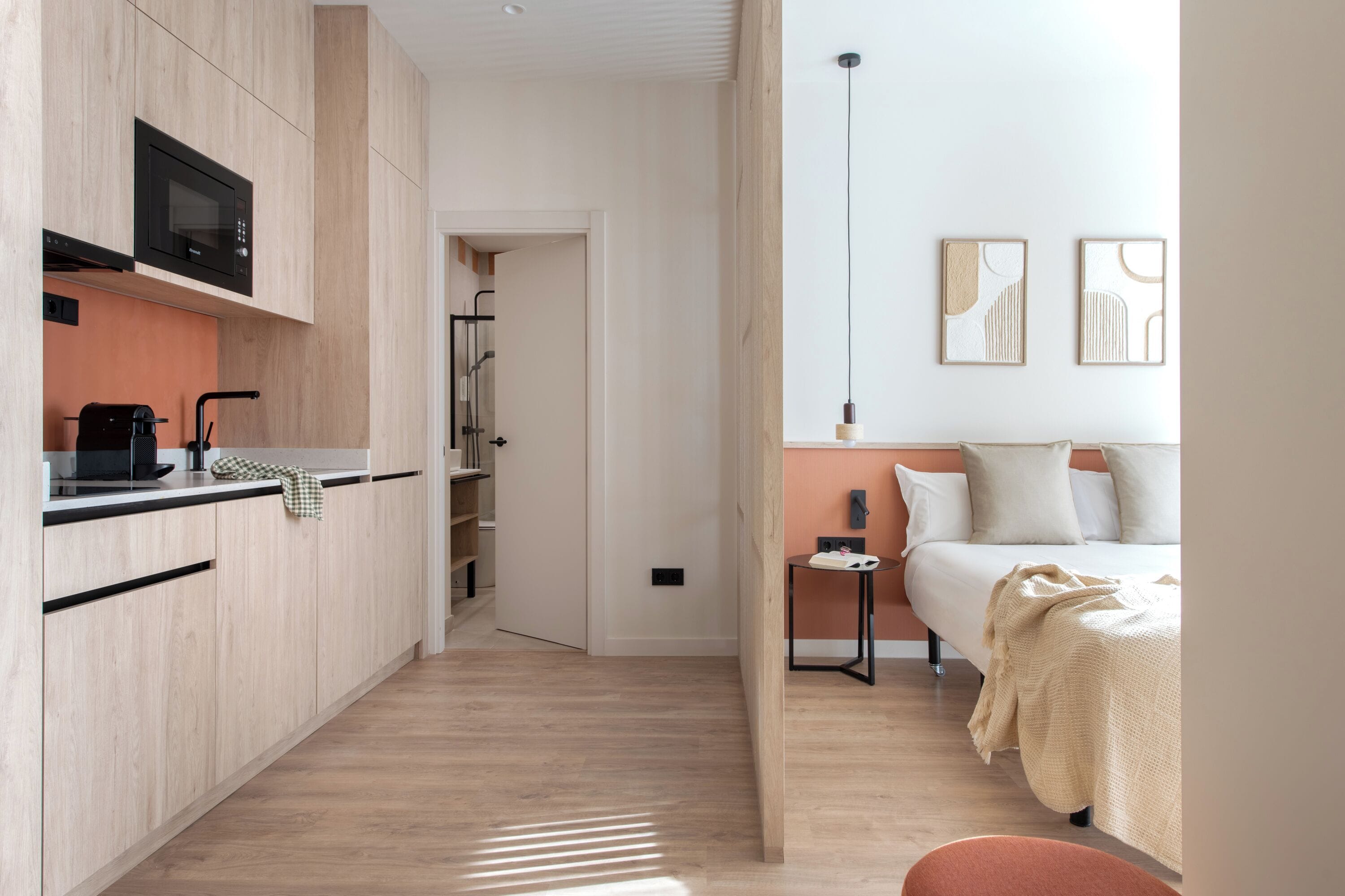 Une chambre d’hôtel moderne et compacte, dotée d’une kitchenette, d’un lit recouvert d’une couverture beige et d’œuvres d’art accrochées au mur.