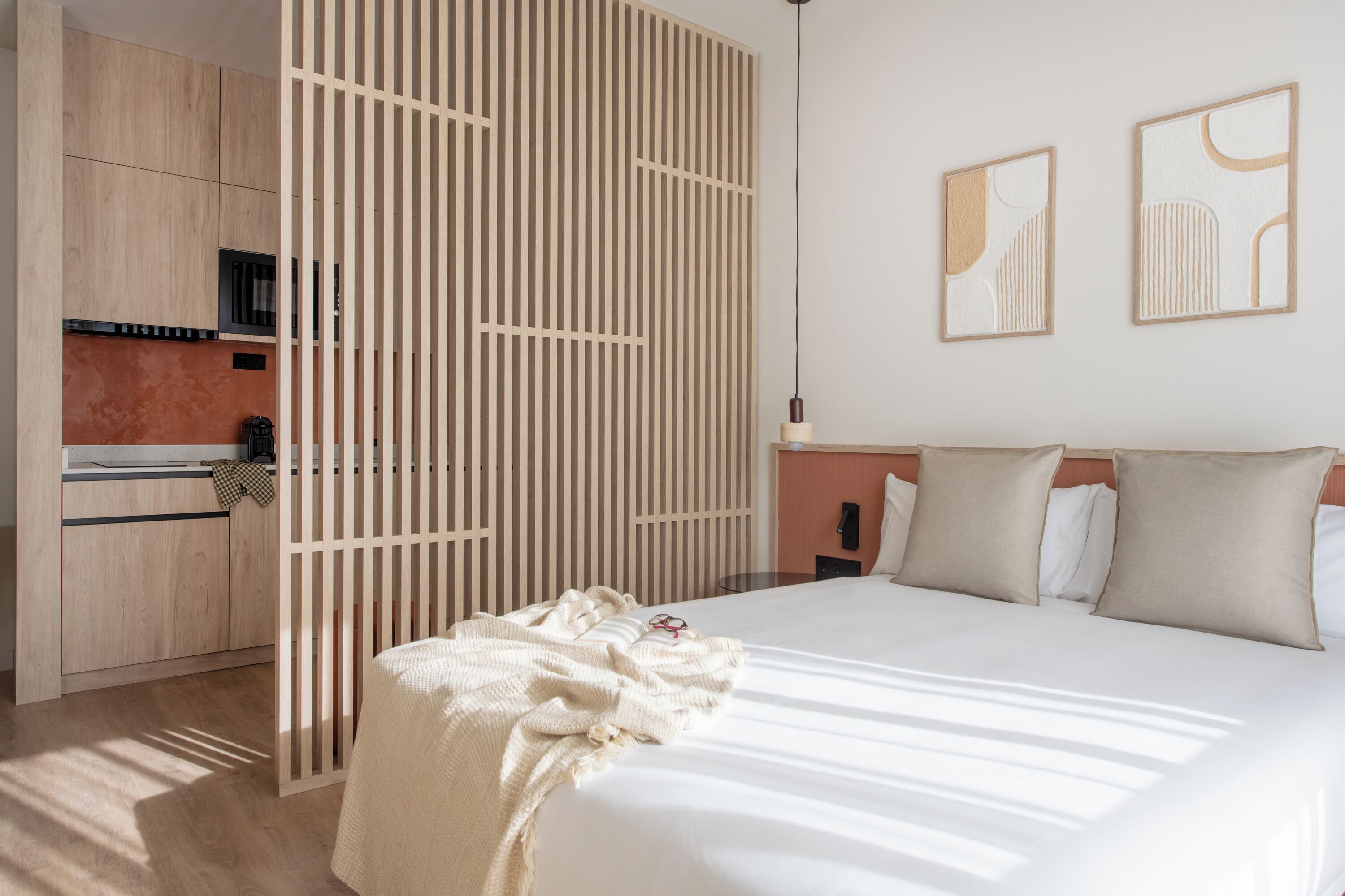 A modern bedroom with a large bed, a wooden partition, a kitchenette, and abstract wall art.