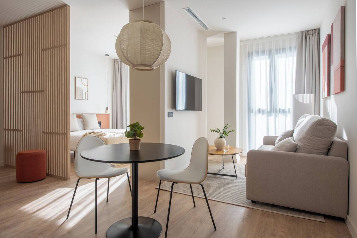 A modern living room with a sofa, a round table with chairs, a TV mounted on the wall, and a large window with curtains.