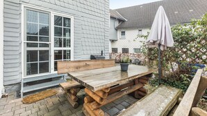 Ein Holztisch mit Bänken, ein Blumentopf und ein überdachter Sonnenschirm vor einem Haus mit weißer Fassade und einem Fenster.