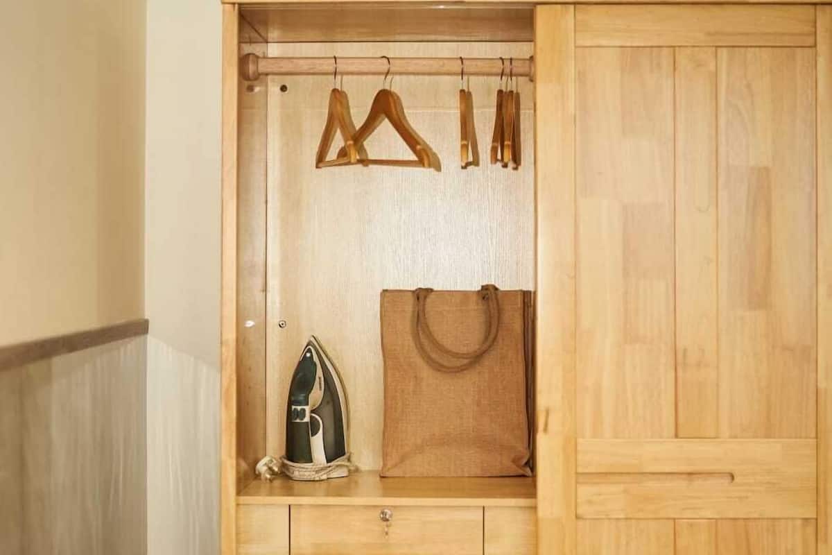 A bedroom with a wooden wardrobe, a hanging clothes rack, an ironing board, and a bag.