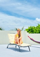 Una persona sentada en una silla reclinable color beige, con una hamaca cerca, al aire libre bajo un cielo despejado y con follaje en el fondo.