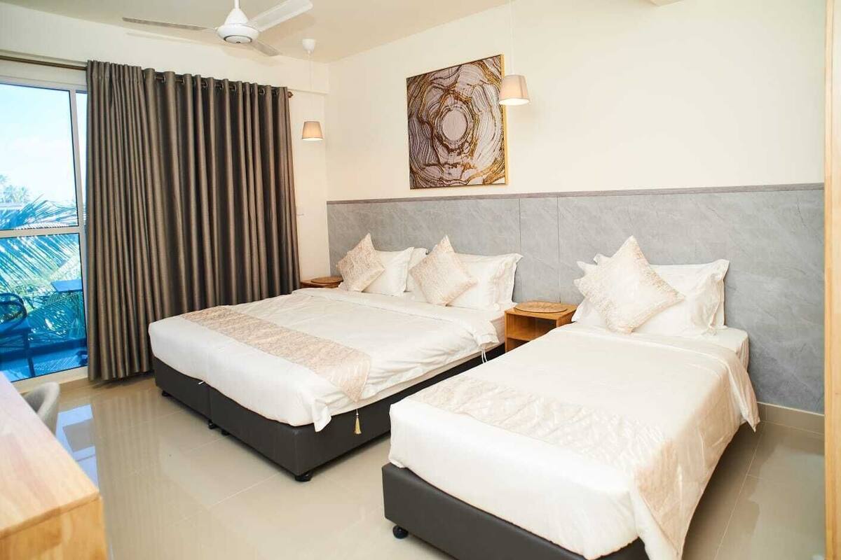 A hotel room with two beds, a large window with curtains, a ceiling fan, a wall-mounted lamp, and a decorative piece above the beds.