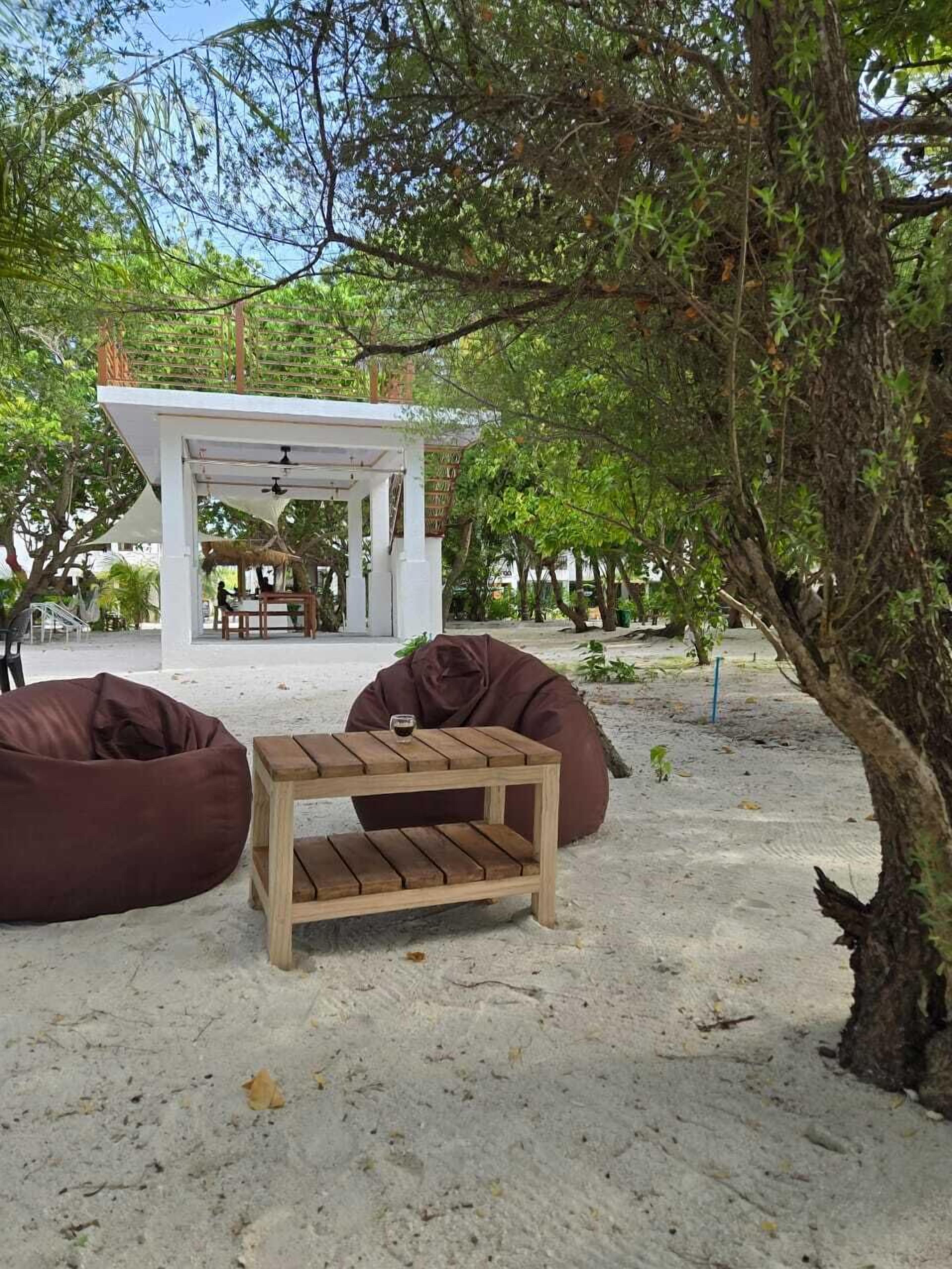 An outdoor seating area with a wooden table and two bean bags, surrounded by trees and a white structure in the background.