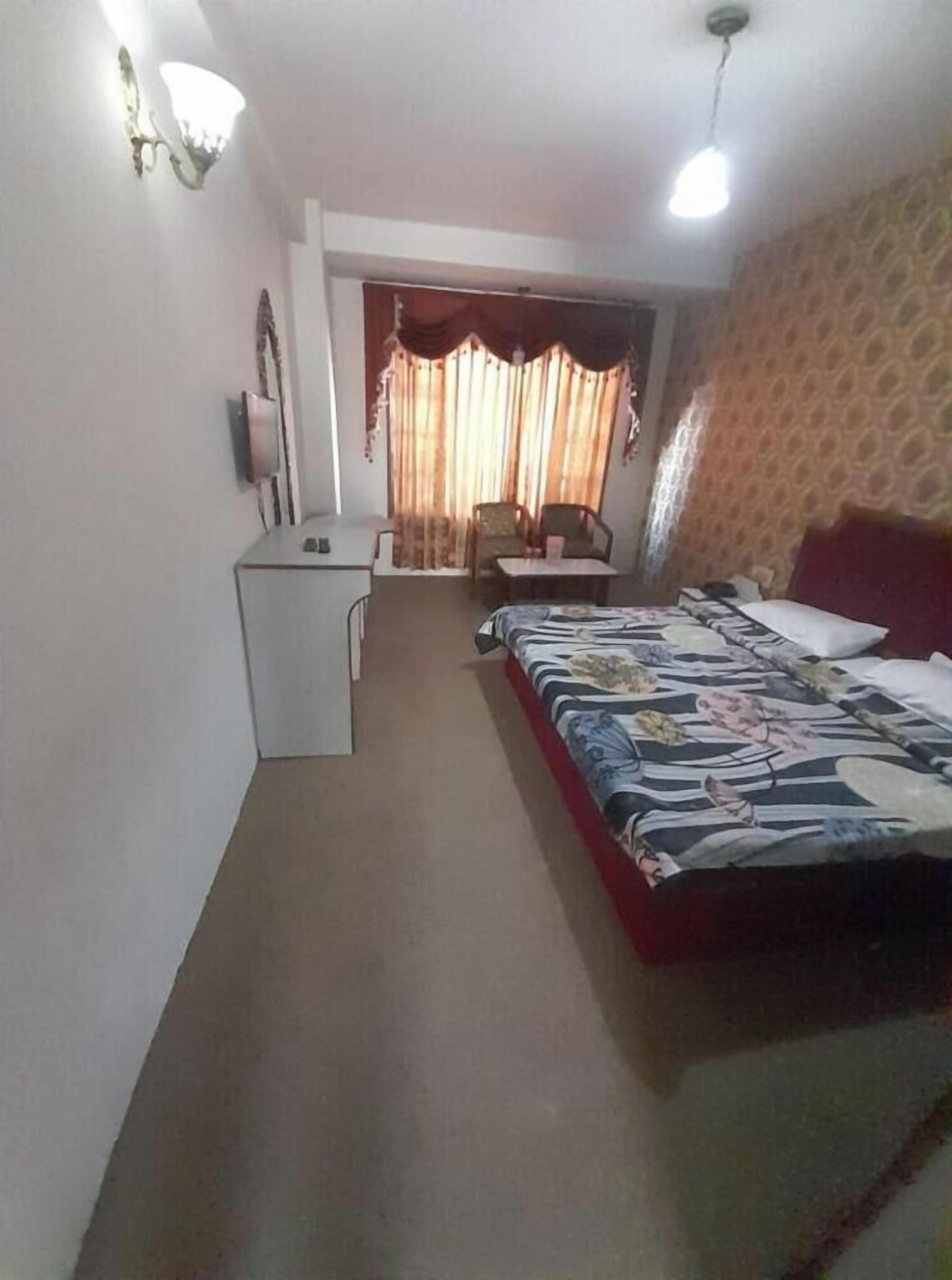 Habitación de hotel con cama, escritorio, silla y una mesita. Se observa una cortina y una pared con diseño.