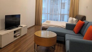 Una habitación de hotel moderna con una televisión de pantalla plana, un sofá con cojines naranjas, una mesa de centro de madera y una cama con sábanas blancas.