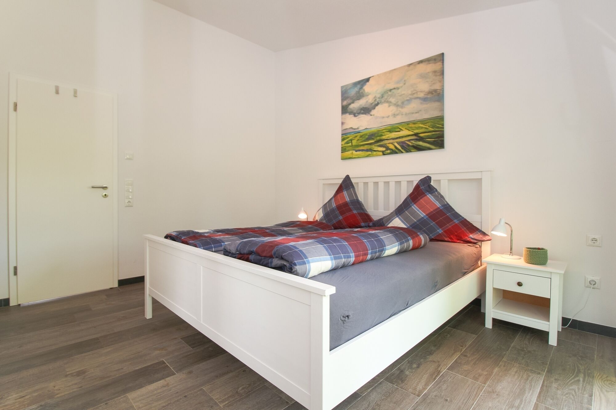 Ein Schlafzimmer mit einem weißen Bett, Karo-Bettwäsche, einem Nachttisch mit Lampe und einem Bild an der Wand.