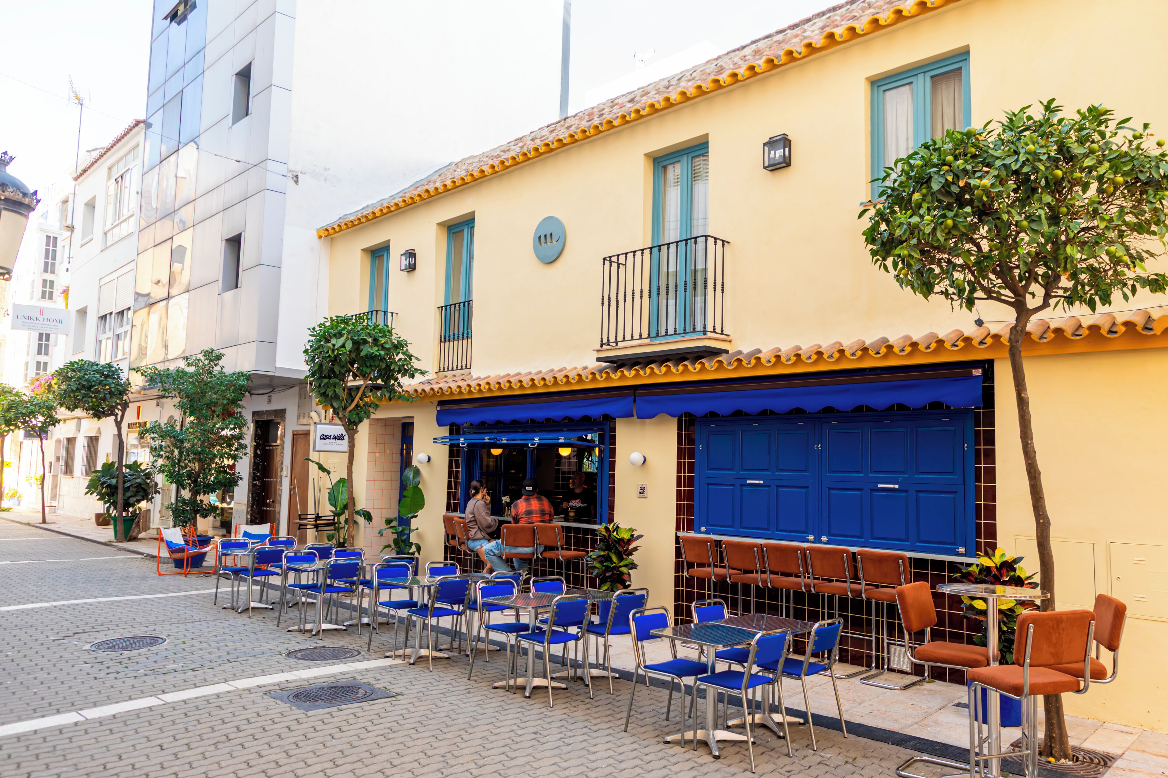 Ein Straßencafé mit blauen und orangen Stühlen, ein gelbes Gebäude mit blauen Fensterläden und ein Baum im Vordergrund.